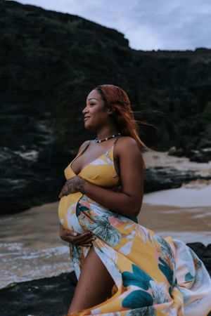 A person in a flowing yellow floral dress poses dramatically against a rocky beach landscape.