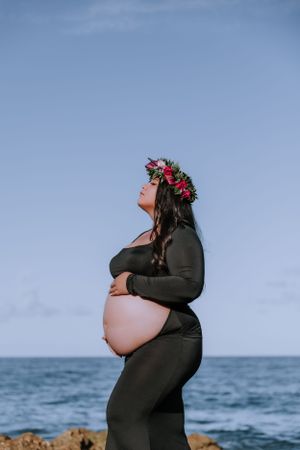 Expectant mother in black dress and flower crown poses against ocean backdrop.