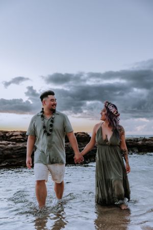 Couple wading in ocean water during sunset with waves and rocks in the background.