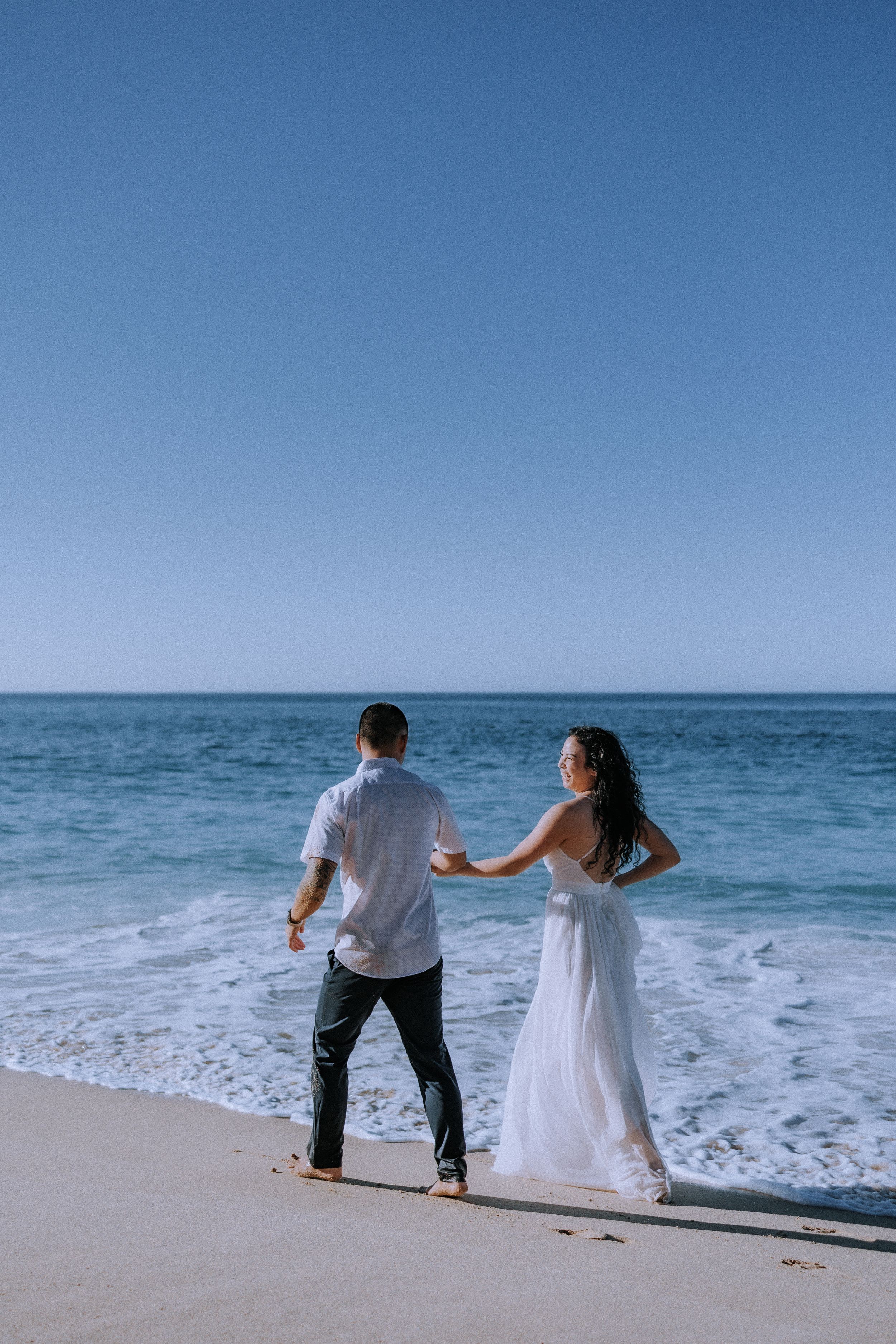 A couple in wedding attire runs playfully along the ocean shoreline on a bright sunny day.