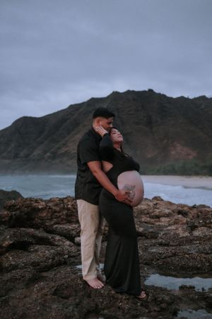 Intimate maternity moment captured on rocky coastline with mountains and ocean views.