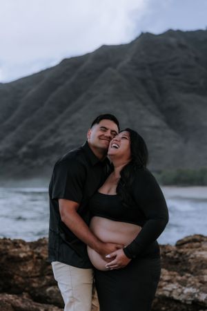 Happy couple sharing joyful moment during maternity photoshoot on Hawaiian coastline.