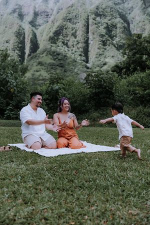 Family relaxing on white blanket in grassy field with dramatic mountain backdrop.