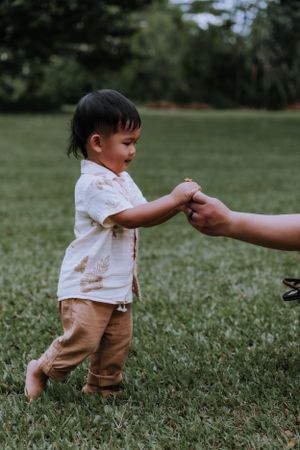 Small child in white shirt and khaki pants holds hands with someone on grassy field.
