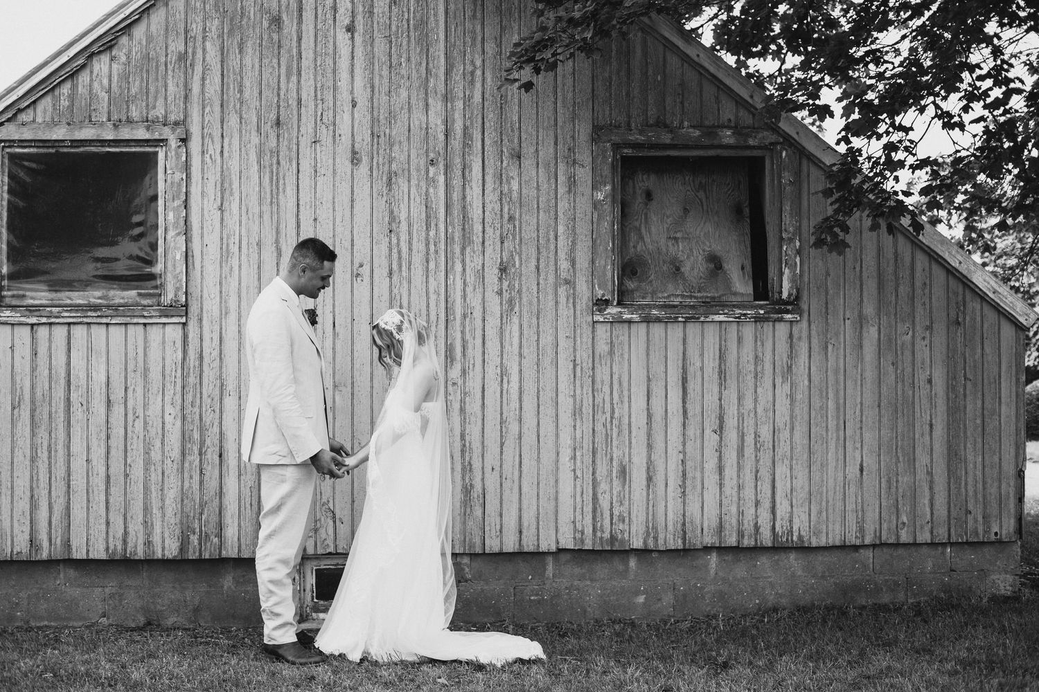 A black and white portrait of newlyweds standing against a rustic wooden barn.
