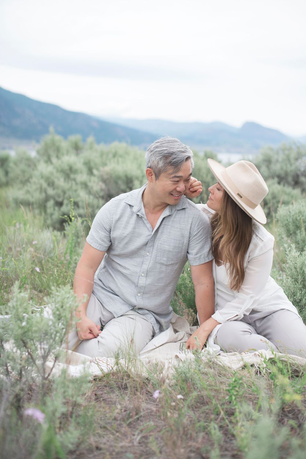 A couple sits intimately together on a hillside surrounded by sage brush and mountain views at Penticton, B.C.