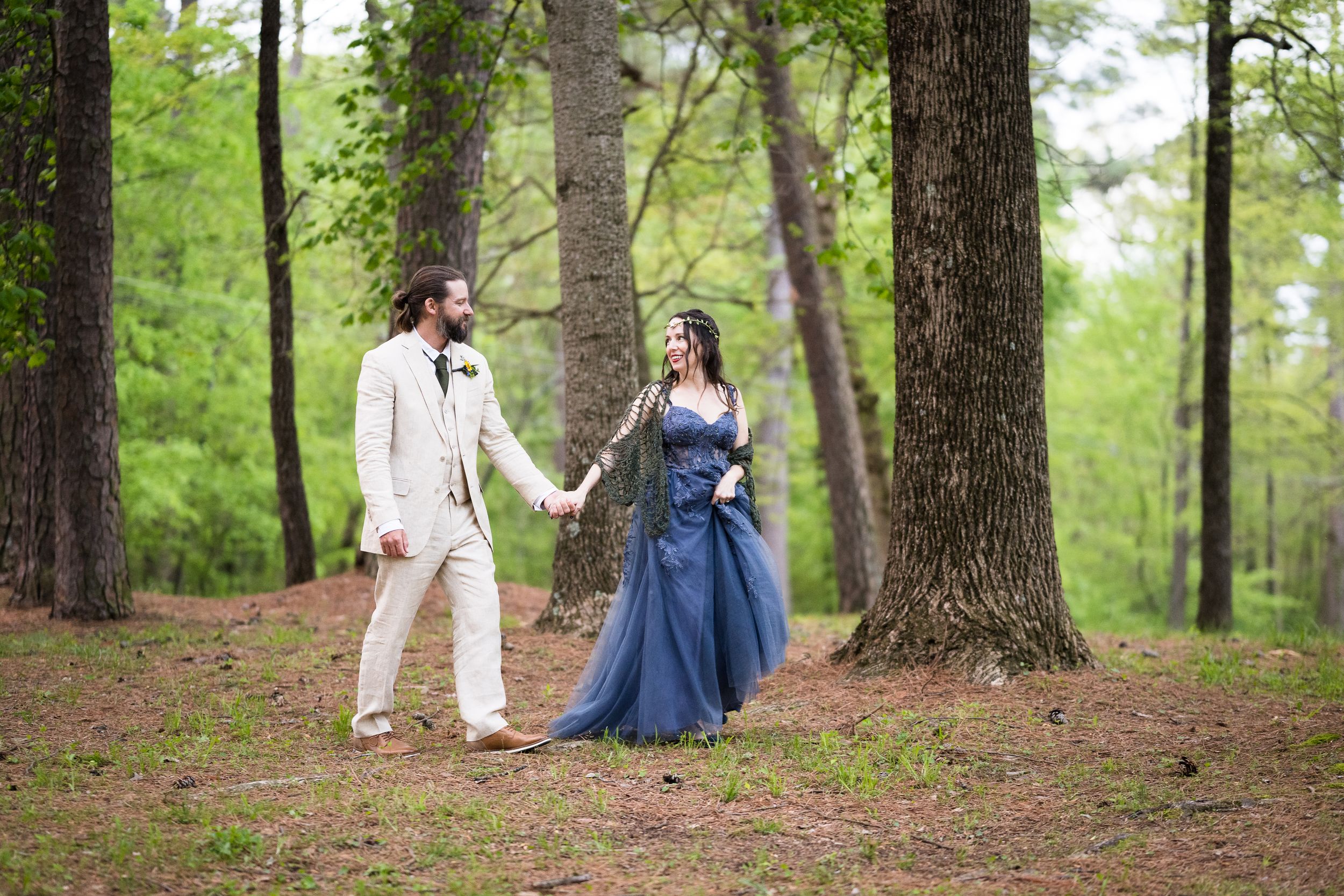 A couple walks hand in hand through a forest wearing formal attire with a blue gown.