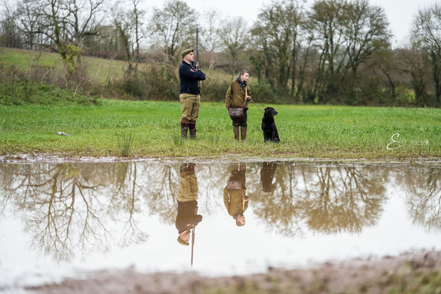 Figures and dog reflected in puddle while standing in green field.