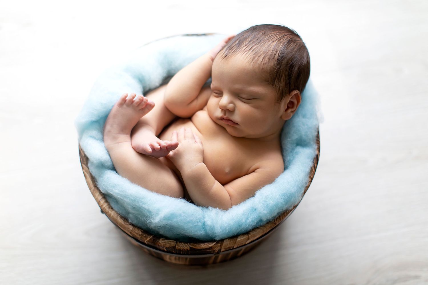 Sleeping newborn baby curled up in blue-lined wooden basket on white background.