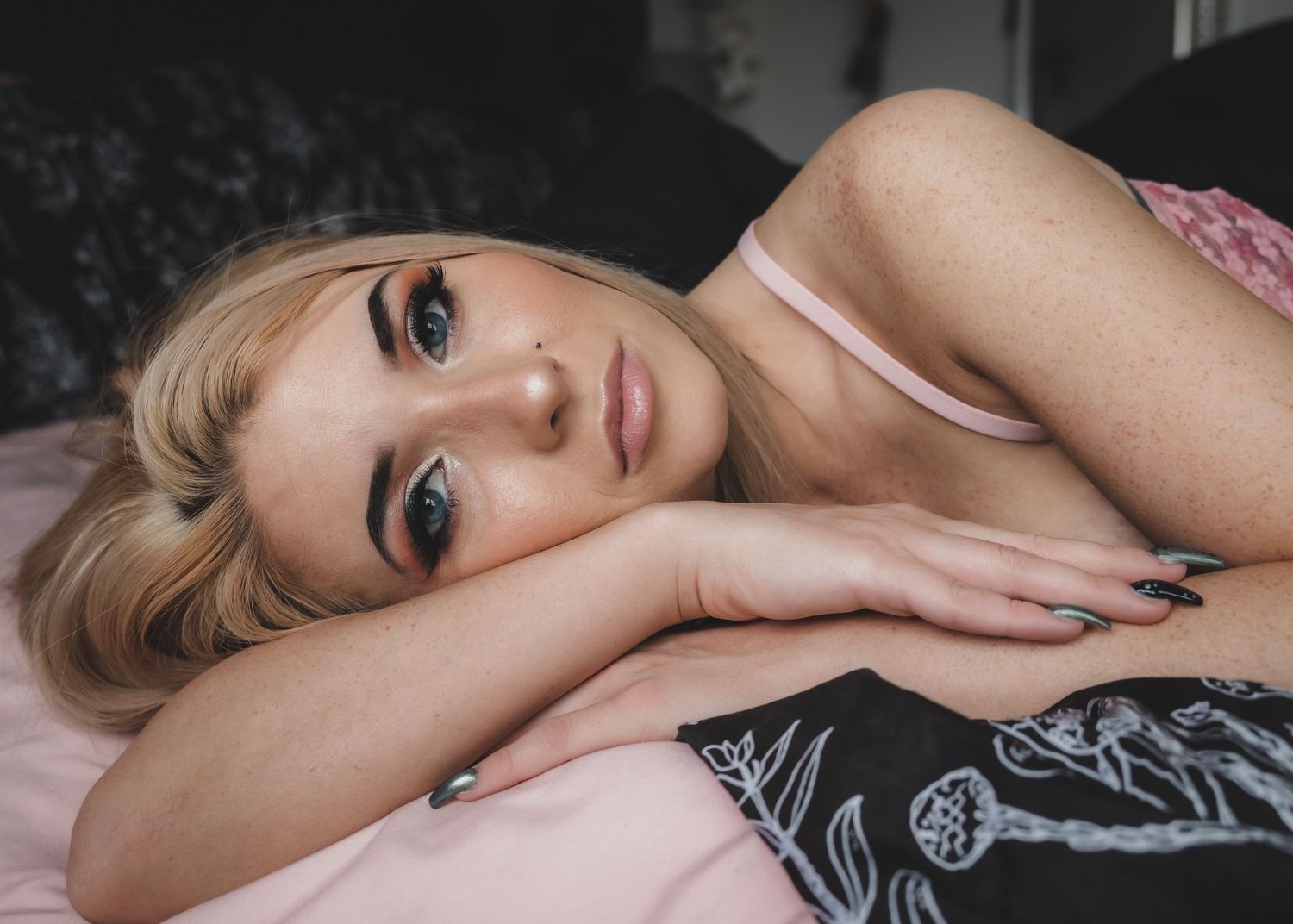 Young blonde woman with blue eyes resting her head on her hands on a pink bed.