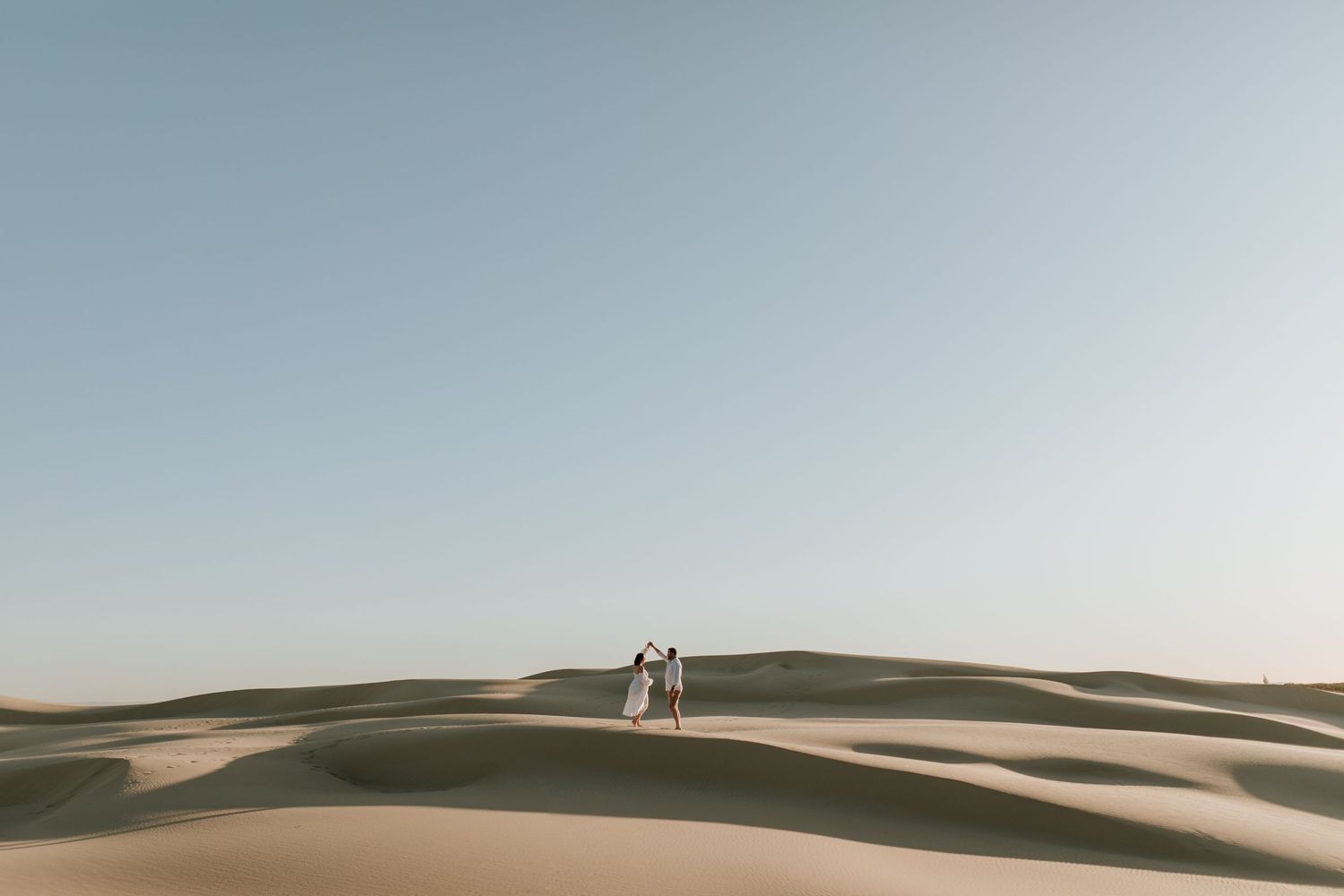 Two silhouettes stand together atop sand dunes in a desert landscape during golden hour.