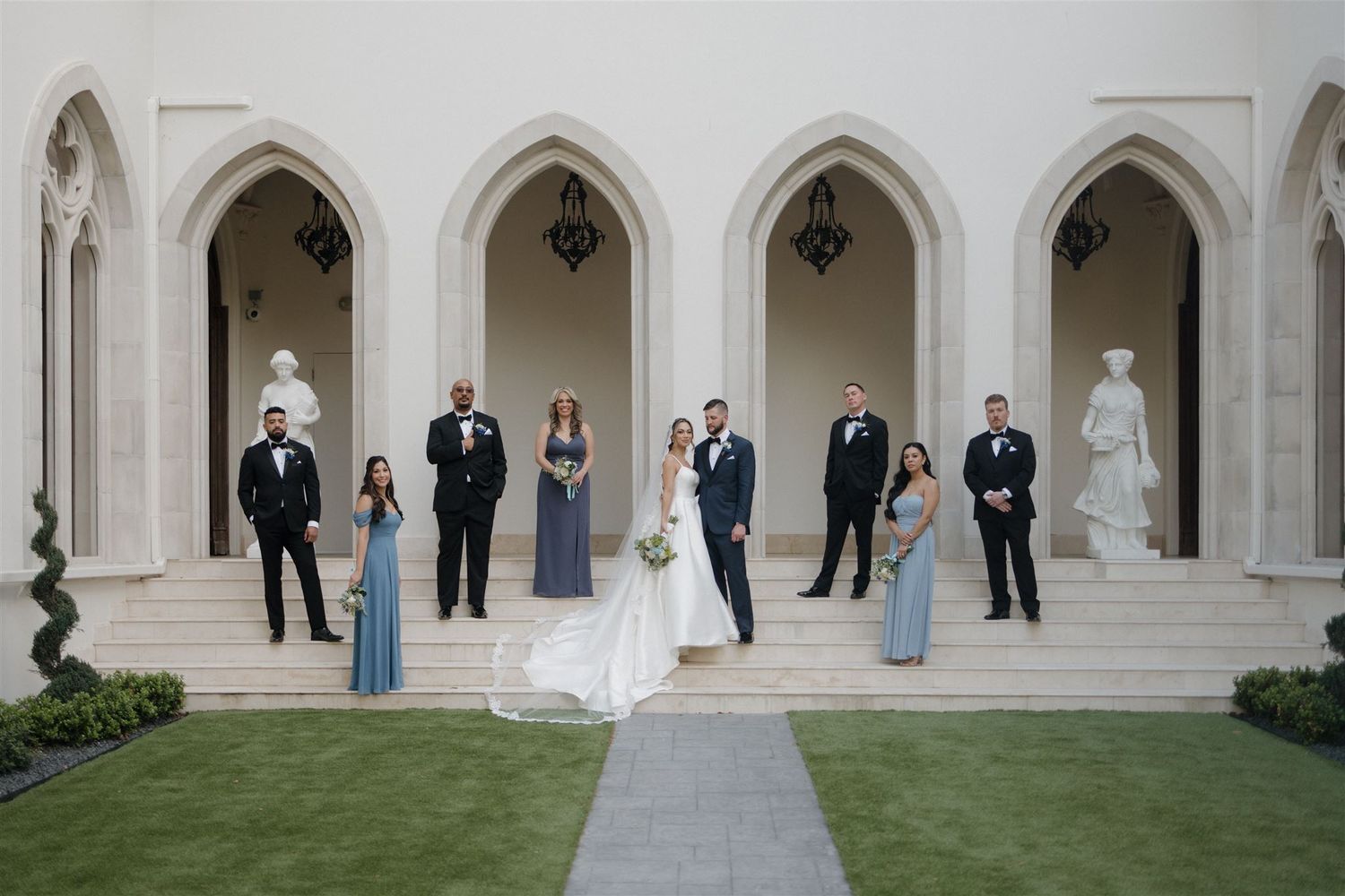 Wedding party members standing in elegant stone archways along a pathway surrounded by manicured grass.