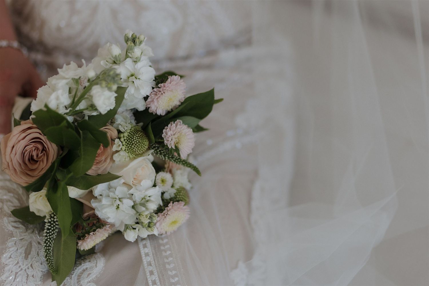 Close up of a bridal bouquet with pink roses, white flowers, and greenery against lace and tulle.
