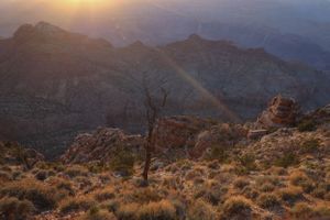 Scenic sunset view over rugged mountain landscape with golden light illuminating rocky terrain and desert brush.