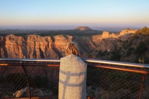 Dramatic overlook of canyon walls with safety railing silhouetted against sunset sky.