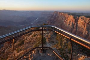 Metal guardrail curves along canyon rim with stunning desert landscape views at sunset.