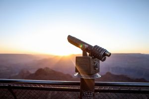 Viewing telescope mounted on observation deck railing overlooking canyon landscape at sunset.