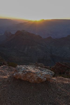 Rocky outcrop overlooking vast canyon terrain with sun setting behind distant mountains.
