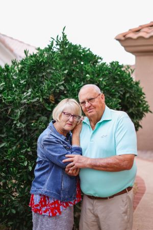 Two people embracing in front of lush green shrubs and a beige stucco wall.