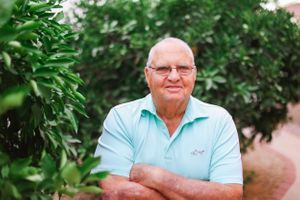 Elderly man in light blue polo shirt sitting outdoors with greenery in background.