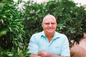 Senior man in casual attire seated outdoors with green foliage behind him.