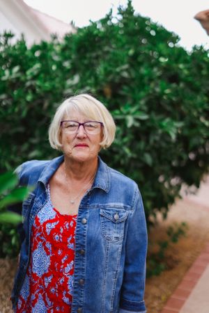 Senior woman in denim jacket and red patterned dress posing in front of green shrubs.