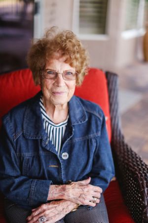A person in a denim jacket and striped shirt sits in a wicker chair with red cushions.