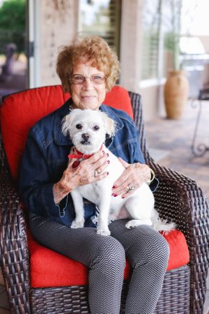 A person holds a small white dog while sitting in a wicker chair with red cushions.