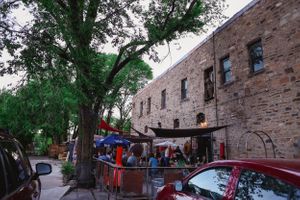 Tree-lined brick building exterior with outdoor seating and parked cars along a street.