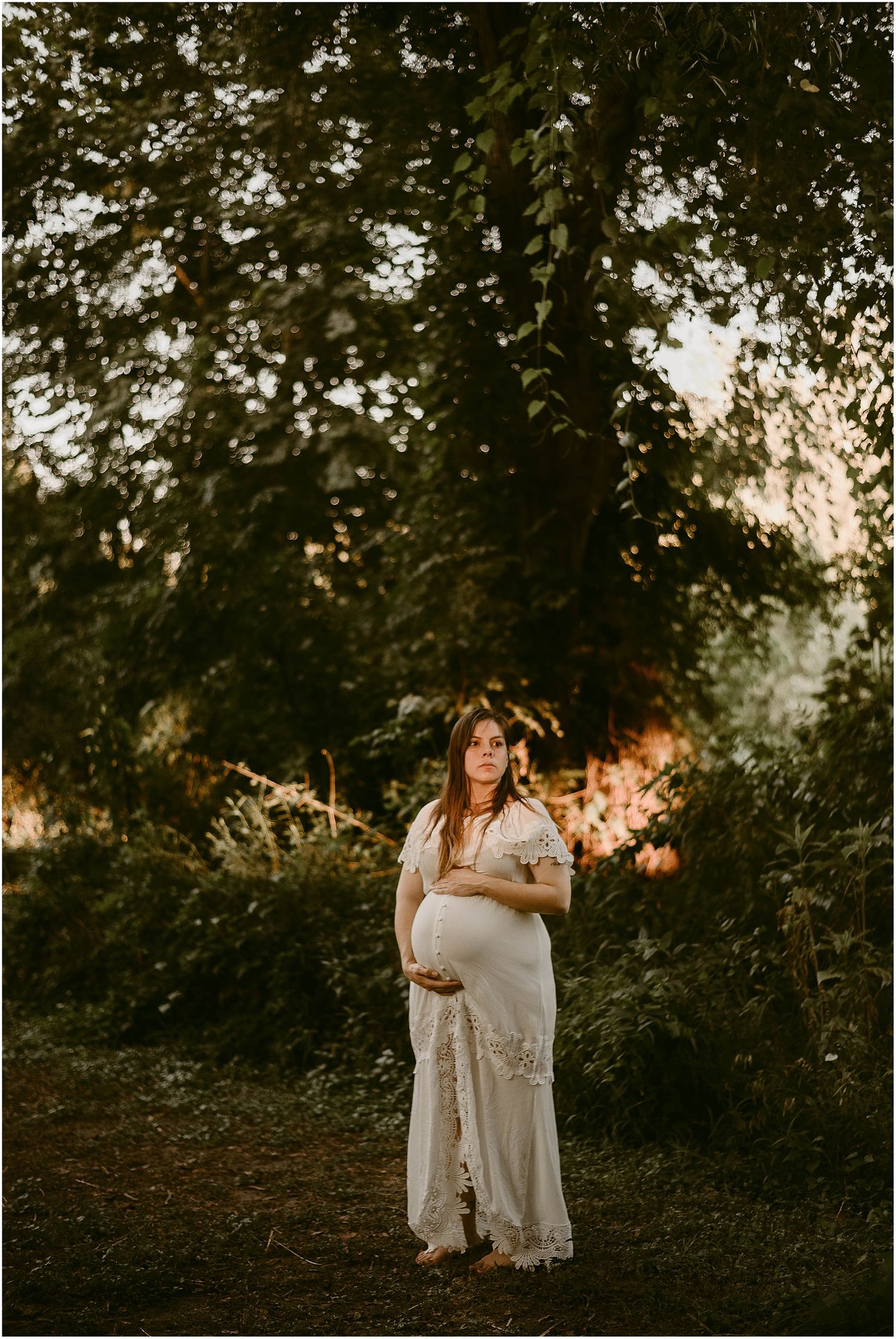 Artistic maternity photograph with dramatic lighting through tree canopy at sunset.