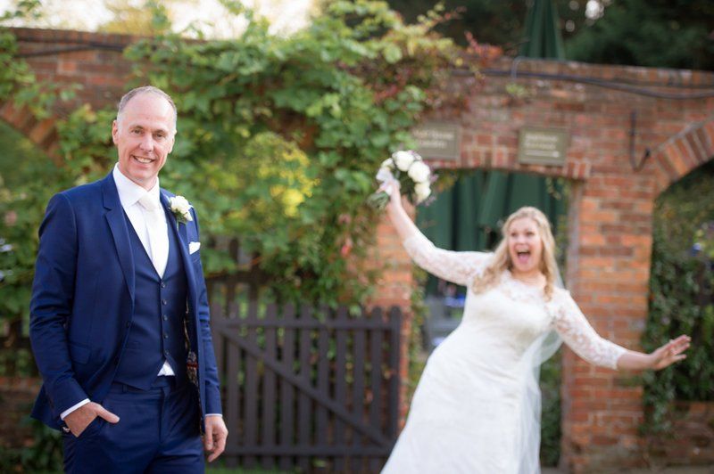Playful wedding couple poses in front of rustic brick wall and garden gate.