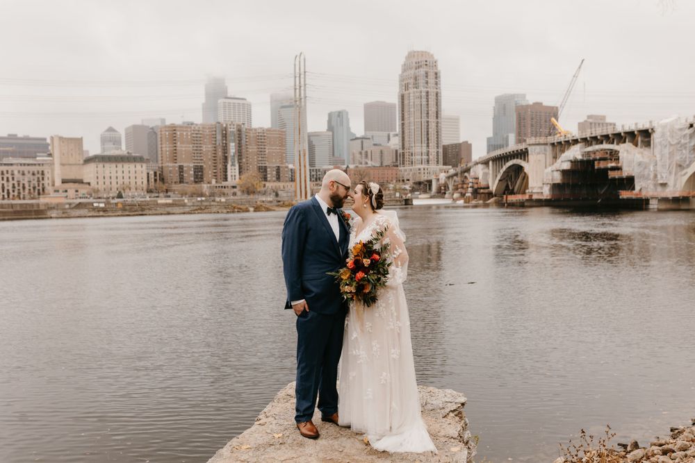 Bride and groom standing close together on rocks by the river with Minneapolis skyline view.