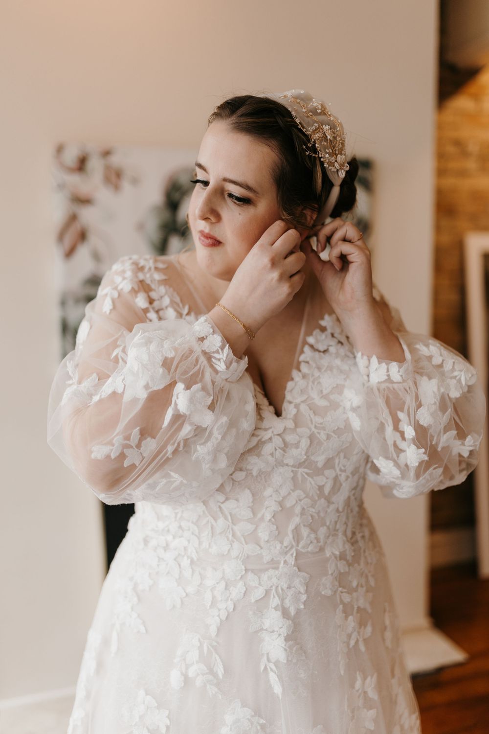 Bride in floral lace gown putting on earrings with gold headpiece in her hair.