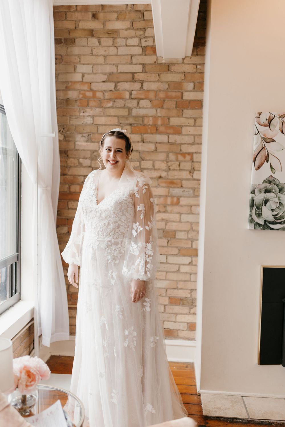 Smiling bride in floral lace long-sleeve wedding gown standing before exposed brick wall.