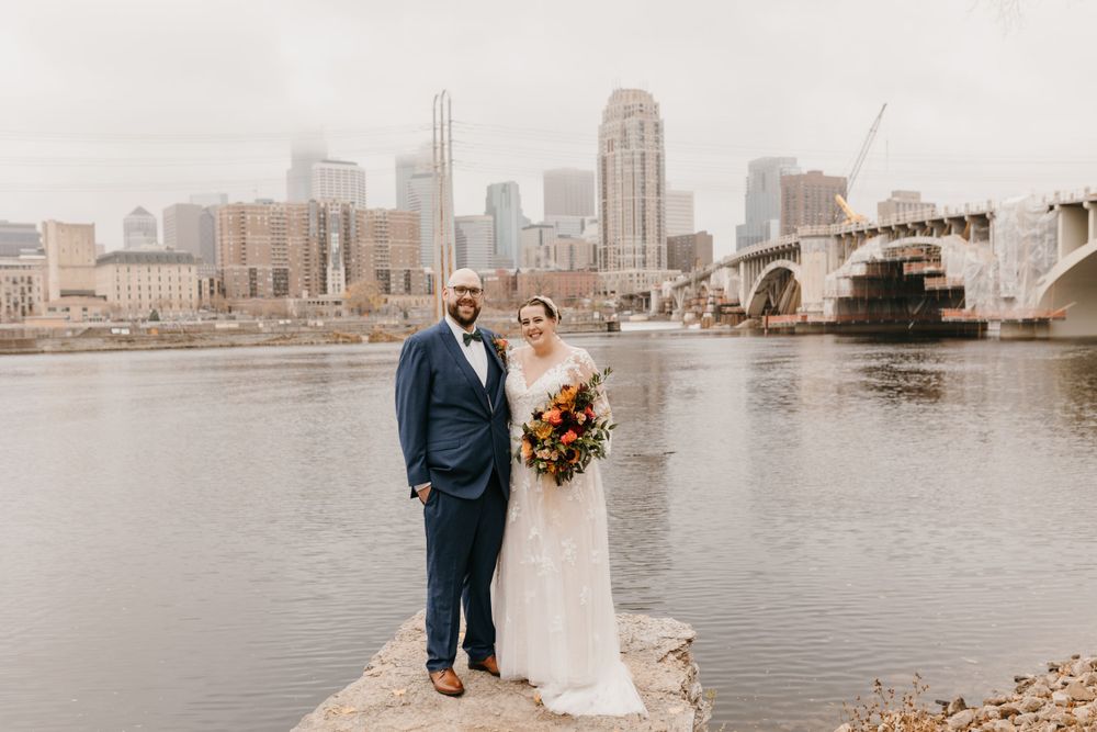 Bride and groom posing by the Mississippi River with Minneapolis skyline and bridge behind them.