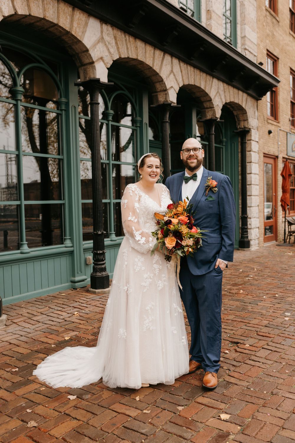 Elegant bride and groom standing together with fall floral bouquet on brick pathway.