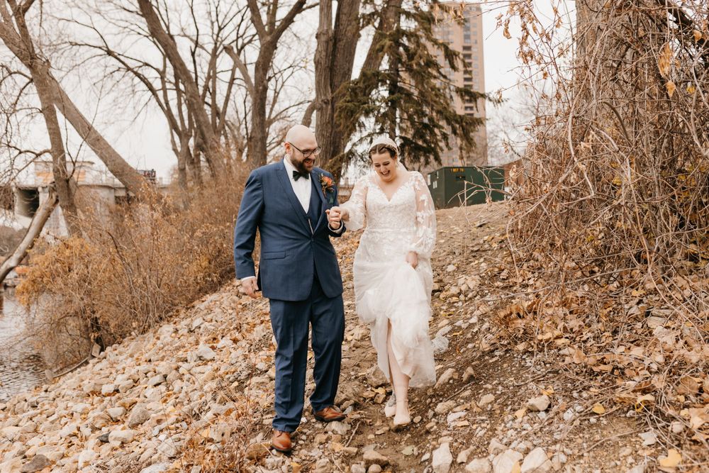 Bride and groom strolling together along a rocky riverbank with bare winter trees.