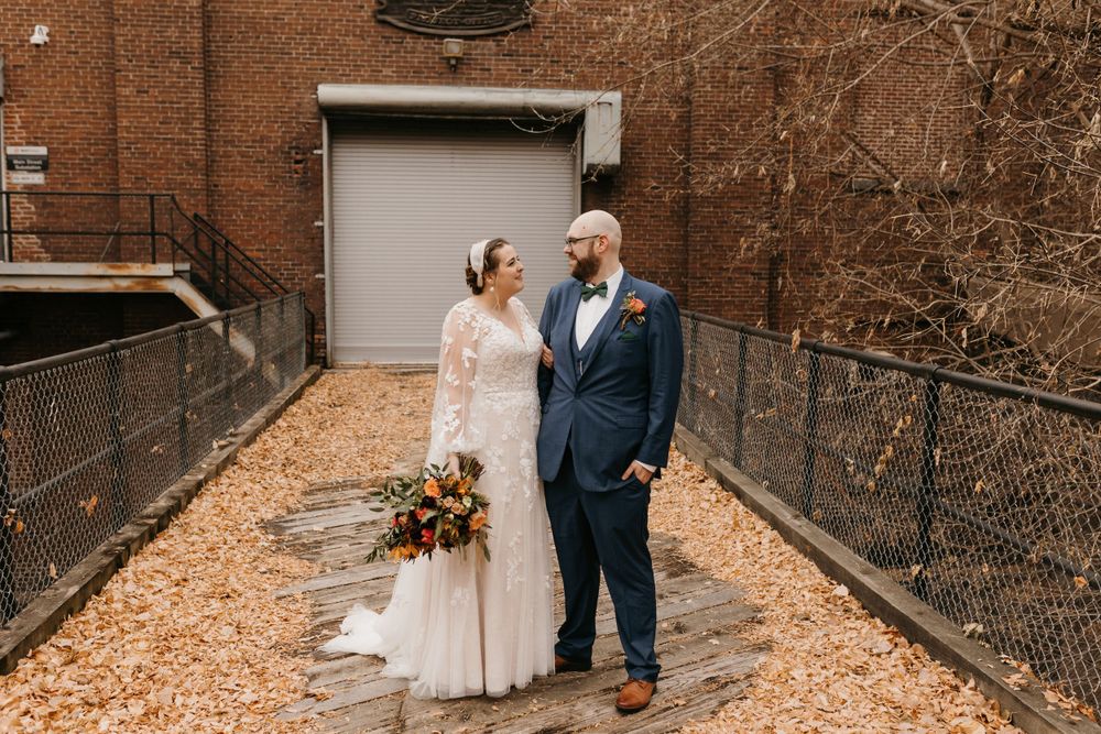 Bride and groom smile at each other on a leaf-covered path beside a brick building.