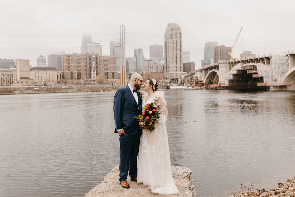 Bride and groom sharing a tender moment by the river with Minneapolis city skyline backdrop.