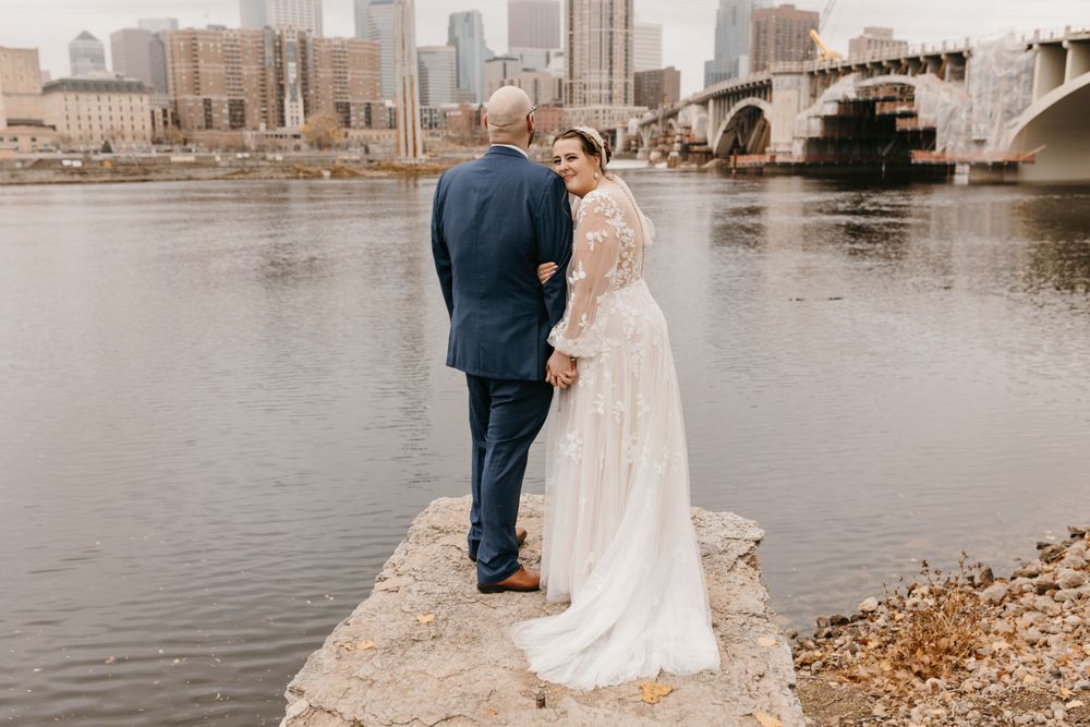 Bride glancing back while holding groom's hand on rocky riverbank with Minneapolis skyline view.