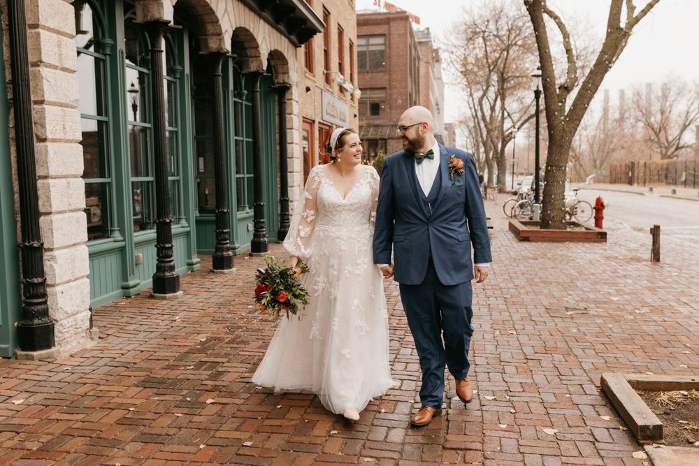 Bride and groom walking hand in hand on brick sidewalk past green-trimmed storefront on their wedding day.