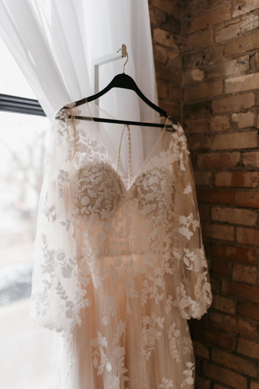 Elegant lace wedding dress with floral embroidery hanging near a brick wall and window.