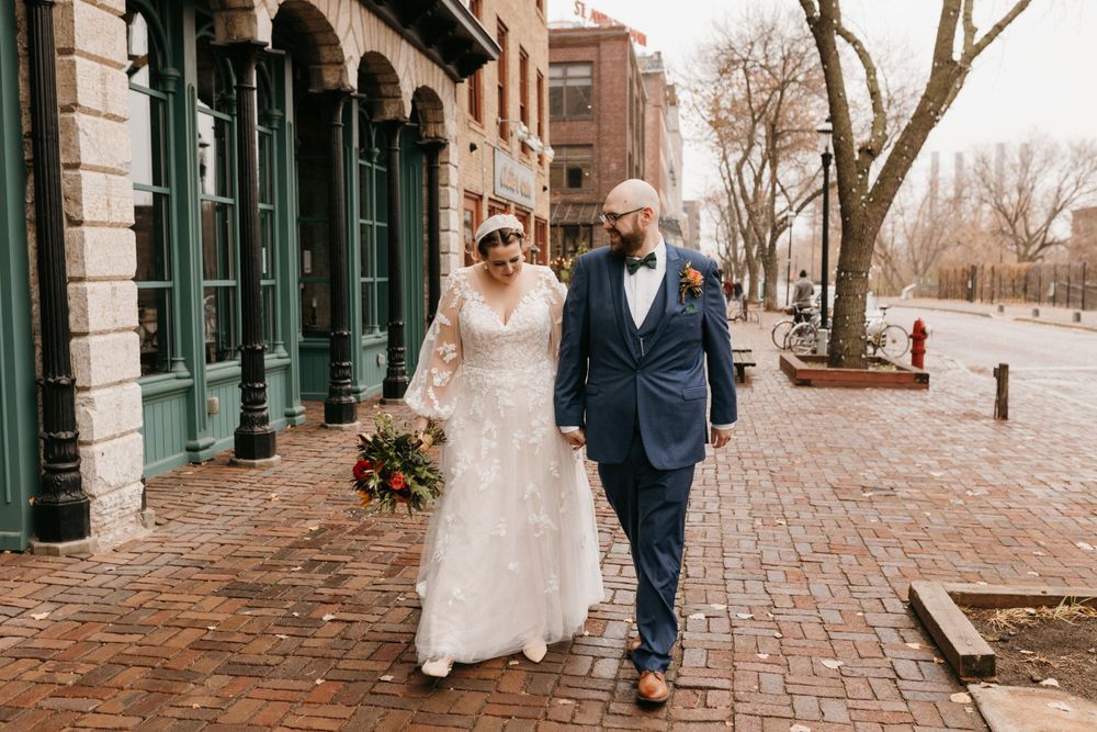 Newlyweds strolling along historic brick street, bride in lace gown holding bouquet, groom in navy suit.