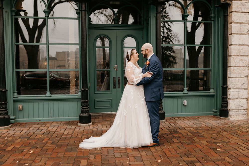Bride and groom embracing tenderly in front of green historic building on wedding day.