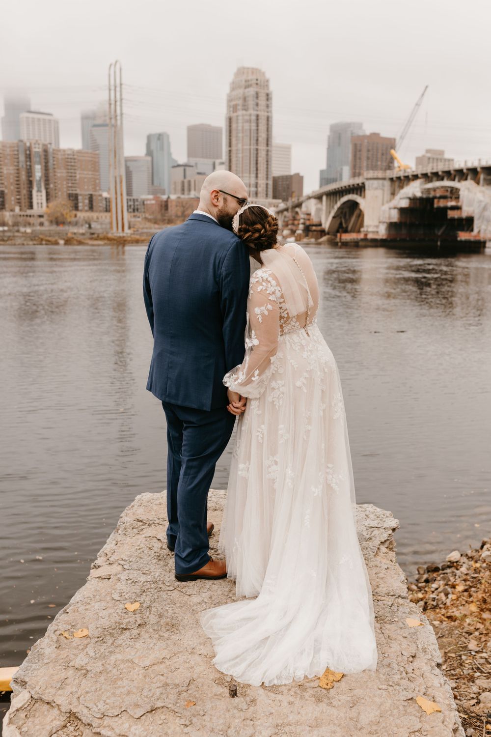 Bride and groom holding hands facing the Minneapolis skyline and bridge from a rocky riverbank.