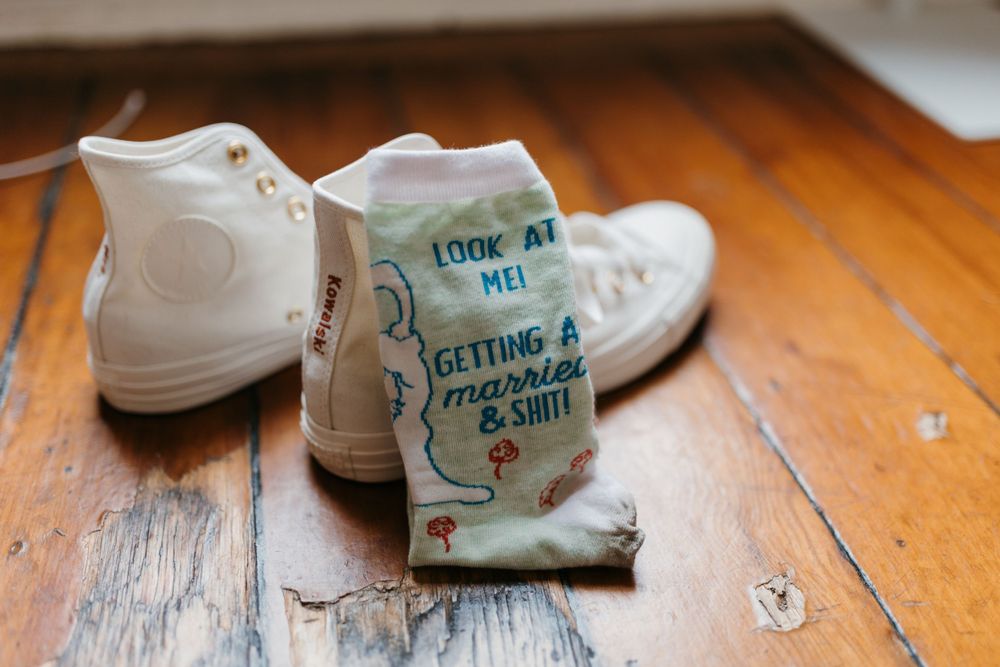 Funny wedding socks reading getting married beside white Converse sneakers on wood floor.