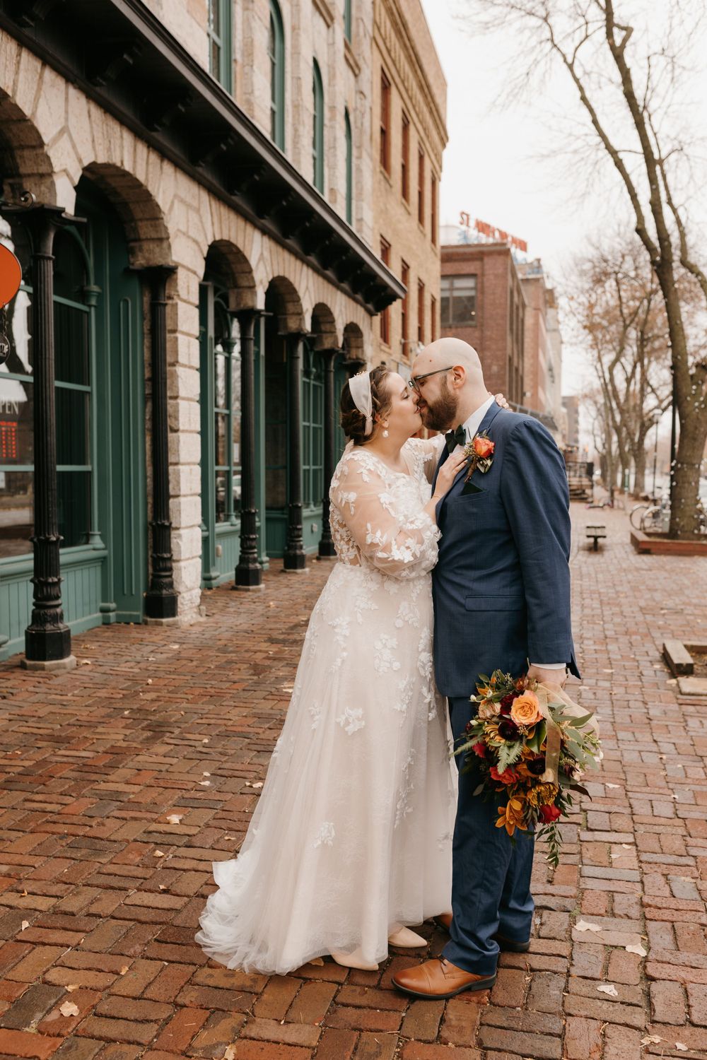 Bride and groom sharing a romantic kiss on brick sidewalk outside historic building on wedding day.