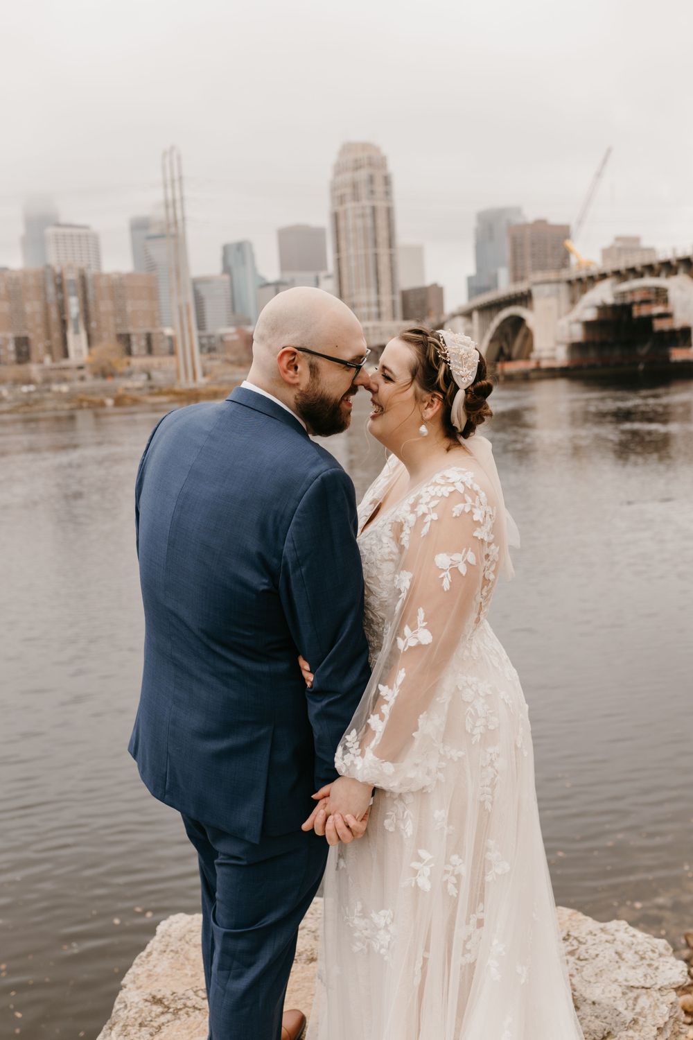 Bride and groom hold hands smiling at each other with Minneapolis skyline and river bridge backdrop.