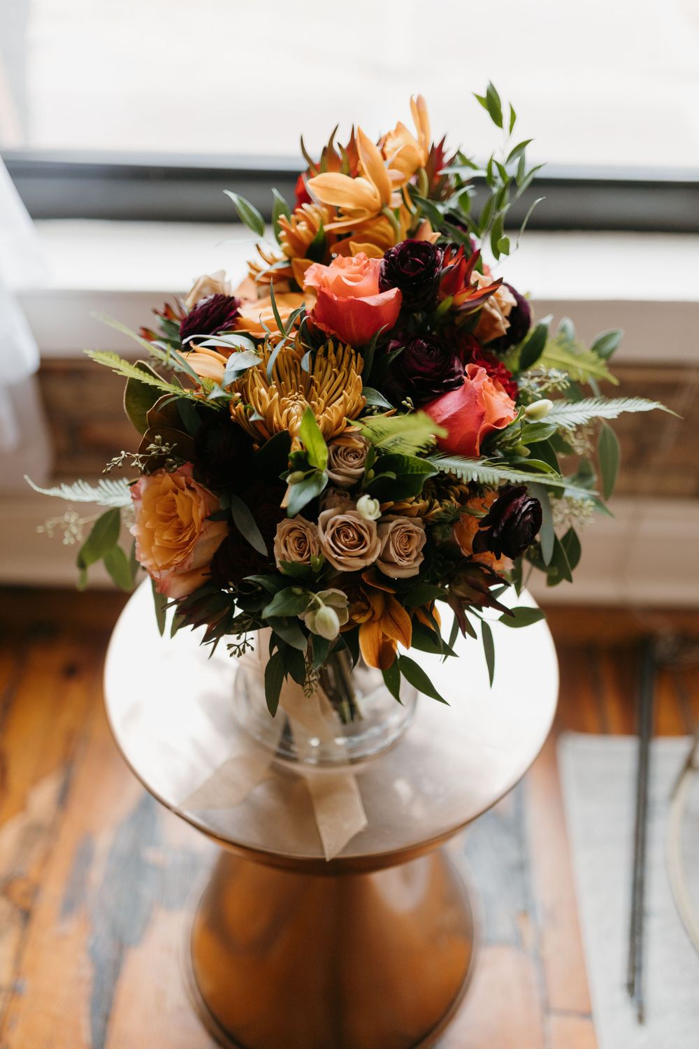 Lush autumn wedding bouquet with orange, red, and burgundy blooms in a glass vase.