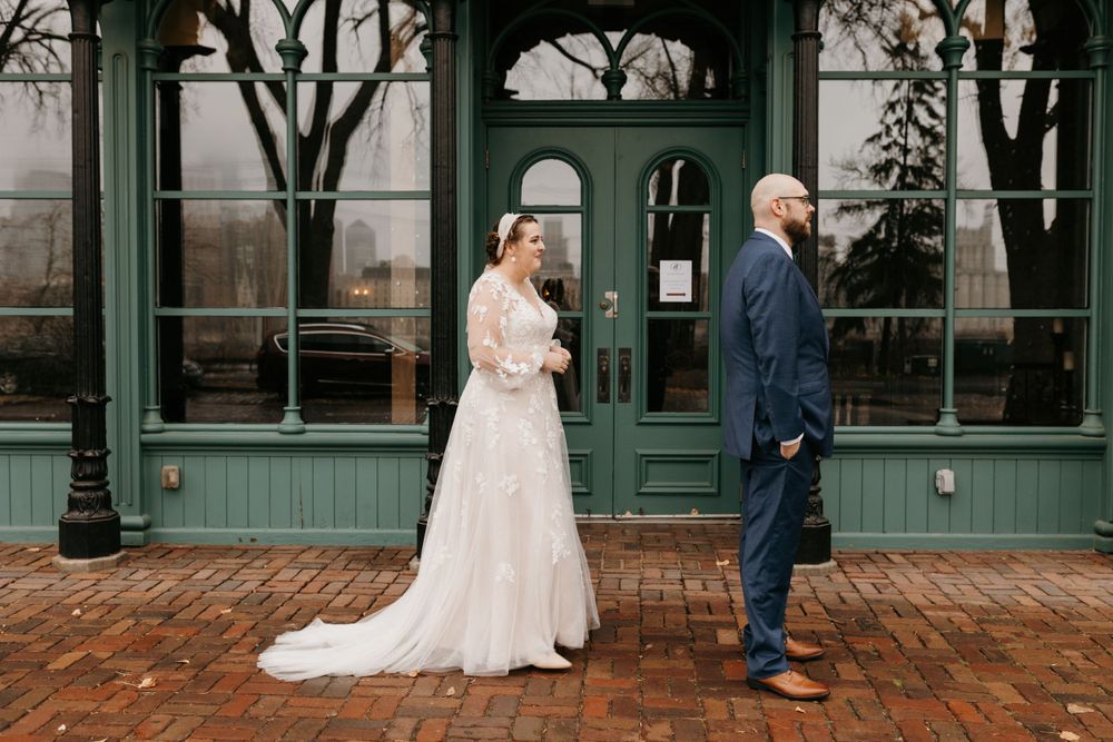 Bride in lace gown approaches groom for first look outside green historic building.